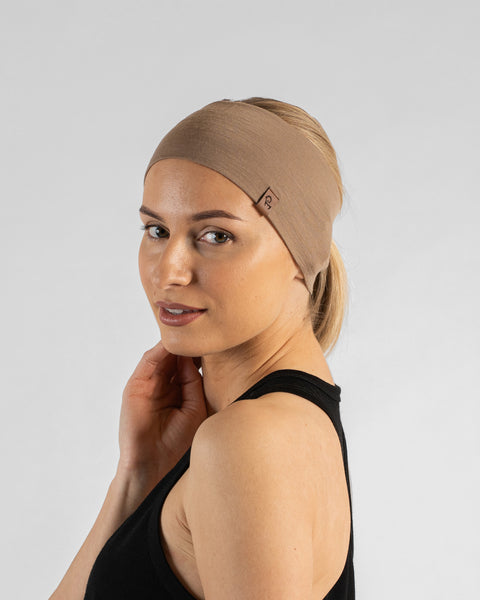A studio portrait of a woman looking over her shoulder directly at the camera with a gentle smile. She is wearing a wide, light beige knit headband that holds back her blonde ponytail, along with a black tank top. A small, square brown leather logo patch is visible on the side of the headband. The background is a solid light grey.