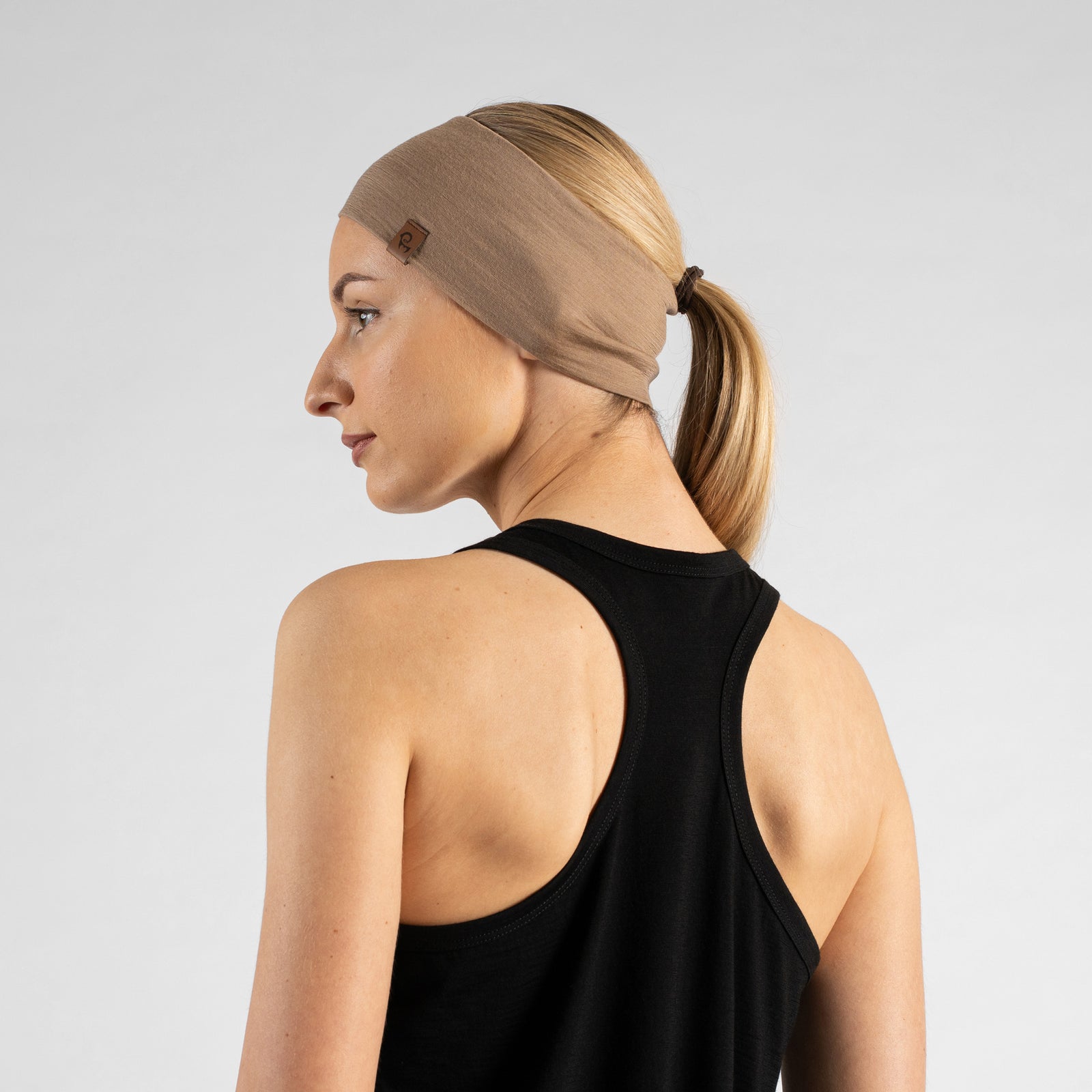A studio portrait of a woman looking over her shoulder directly at the camera with a gentle smile. She is wearing a wide, light beige knit headband that holds back her blonde ponytail, along with a black tank top. A small, square brown leather logo patch is visible on the side of the headband. The background is a solid light grey.