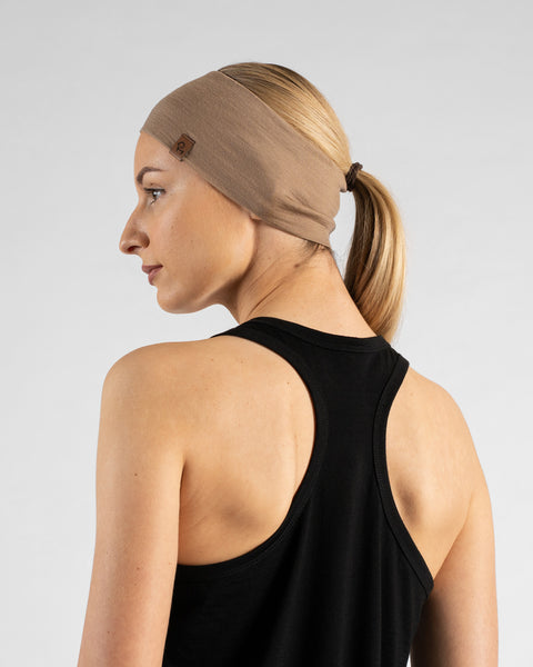 A studio portrait showing the side profile and back of a blonde woman. She is wearing a black sleeveless racerback sport top and a wide beige headband over her hair, which is tied in a ponytail. The background is a plain light grey.
