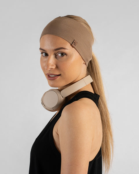 A studio portrait of a blonde woman looking back over her left shoulder at the camera with a gentle smile. She is wearing a black sleeveless sport top, a wide beige headband over her ponytail, and beige headphones resting around her neck.