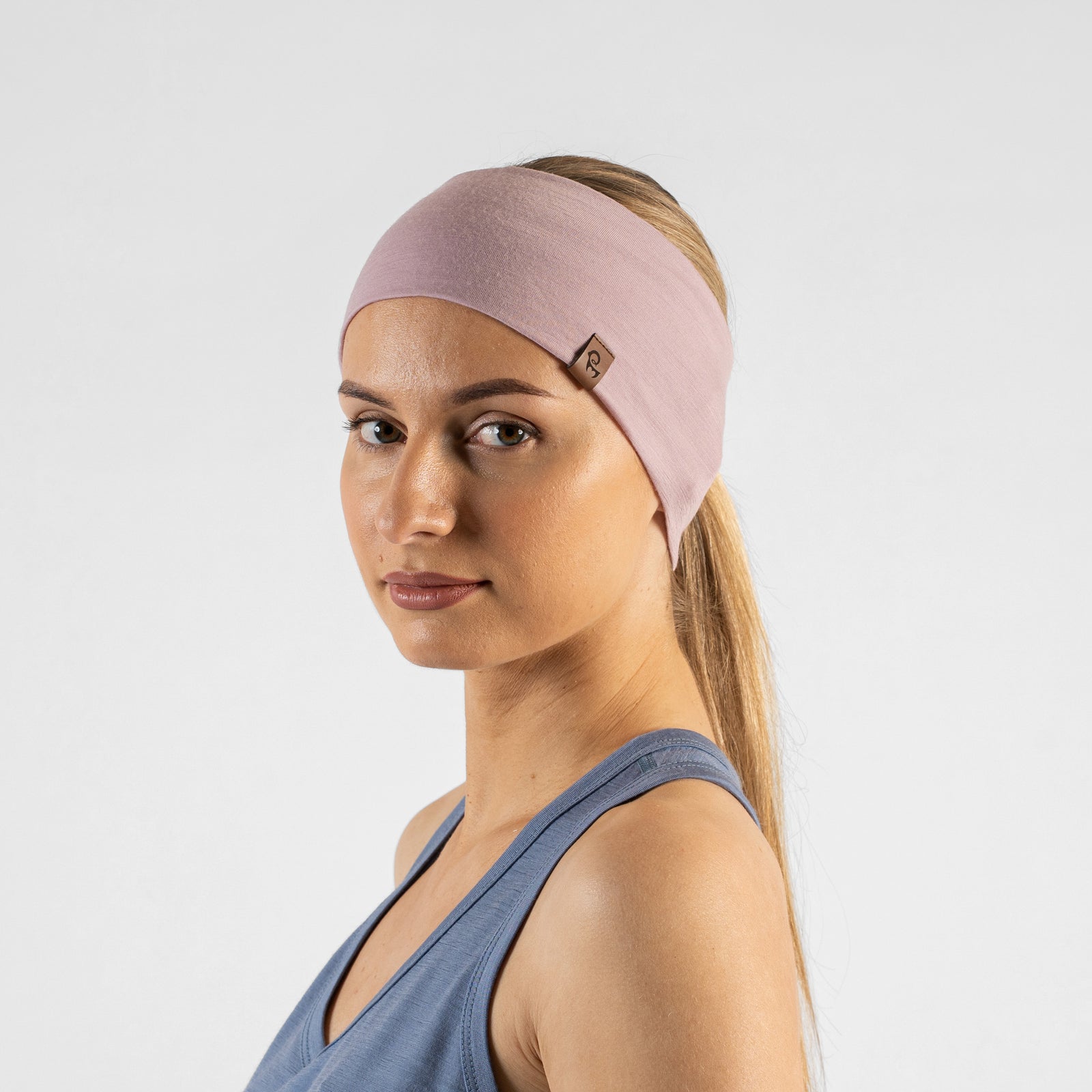 A studio portrait of a blonde woman looking downward and to the left. She is wearing a dusty blue sleeveless top and a dusty pink headband, with both hands raised behind her head to adjust the band against a light grey background.