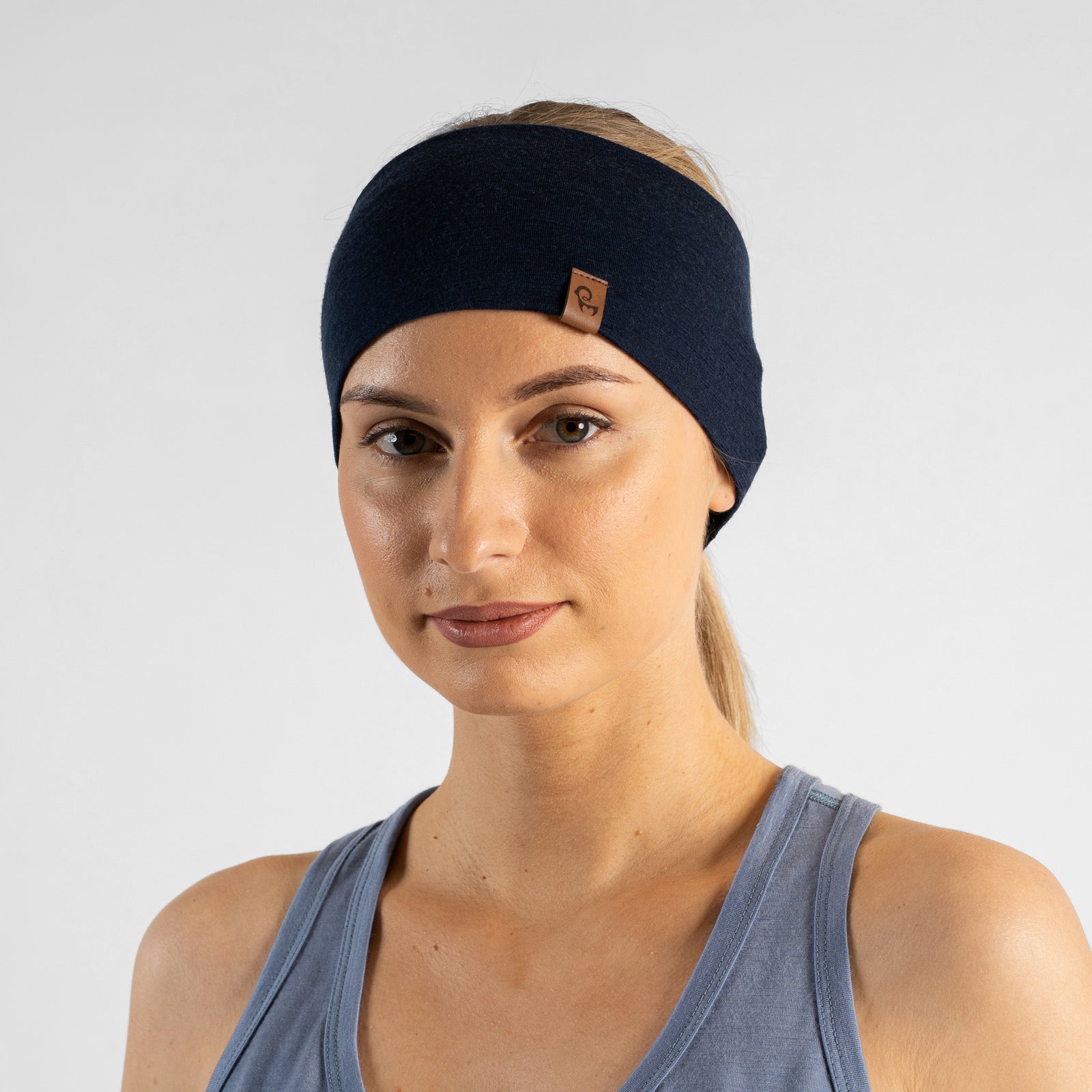 A studio portrait of a blonde woman looking down and off to the left side of the frame against a plain light grey background. She is wearing a dusty blue v-neck sleeveless top and a wide navy blue headband with a small brown logo tag.