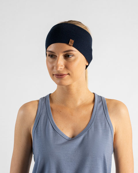 A studio portrait of a blonde woman looking down and off to the left side of the frame against a plain light grey background. She is wearing a dusty blue v-neck sleeveless top and a wide navy blue headband with a small brown logo tag.