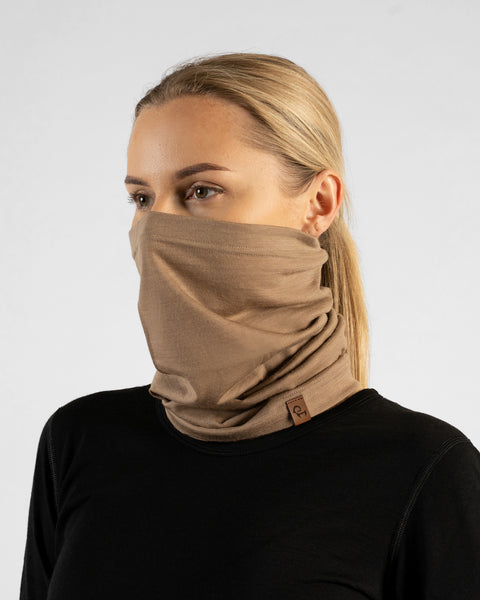 A studio portrait of a blonde woman looking off to the left side of the frame. She is wearing a light beige neck gaiter pulled up to cover her nose and lower face, along with a solid black long-sleeved top. A small brown leather logo patch is visible on the bottom edge of the gaiter. The background is a plain light grey.