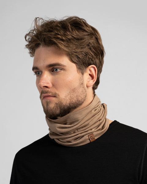 A studio portrait of a man looking off to the left. He is wearing a black long-sleeved shirt and a beige neck gaiter bunched loosely around his neck against a light grey background.