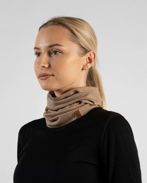 A studio portrait of a blonde woman looking off to the left against a plain light grey background. She is wearing a black long-sleeved shirt and a beige neck gaiter scrunched down around her neck, featuring a small brown logo tag.