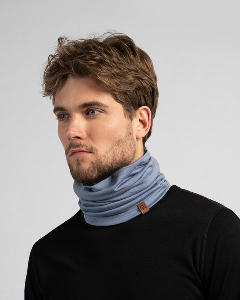 A studio portrait of a man looking off to the left side of the frame. He is wearing a black long-sleeved shirt and a dusty blue neck gaiter bunched around his neck, featuring a small brown logo tag.