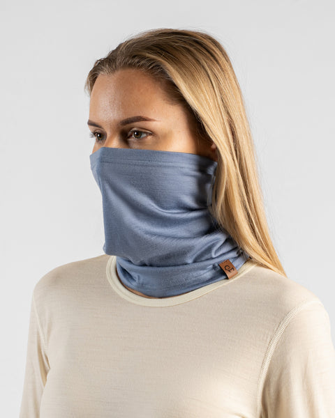 A studio portrait of a blonde woman looking off to the left side of the frame. She is wearing a dusty blue neck gaiter pulled up to cover her nose and lower face, paired with a light cream, natural-colored long-sleeved top. A small brown leather logo patch is visible on the bottom edge of the gaiter. The background is a plain light grey.
