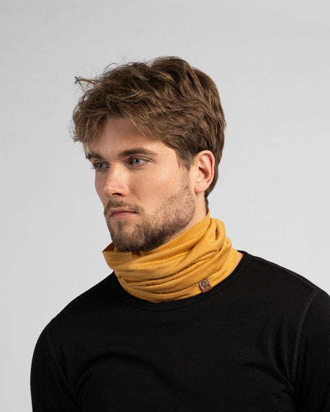 A studio portrait of a man looking to the left against a light grey background. He wears a black long-sleeved shirt and a mustard yellow neck gaiter bunched around his neck.