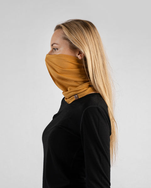 A side profile studio portrait of a blonde woman facing left against a plain light grey background. She is wearing a black long-sleeved shirt and a mustard yellow neck gaiter pulled up to cover her nose and mouth, with a small brown logo tag visible at the base.