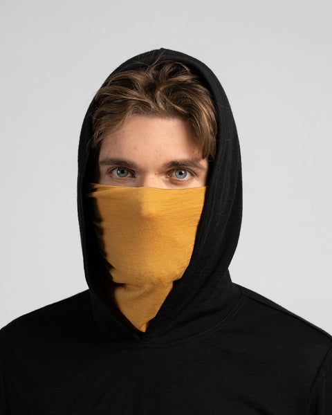 A studio portrait of a man looking directly at the camera, wearing a black hooded shirt with the hood pulled up. His nose and mouth are covered by a mustard yellow neck gaiter.