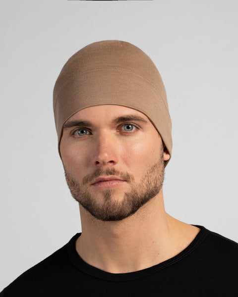 A close-up studio portrait of a man looking directly at the camera. He is wearing a fitted, light beige skull cap and a solid black crew-neck top. The background is a plain light grey.