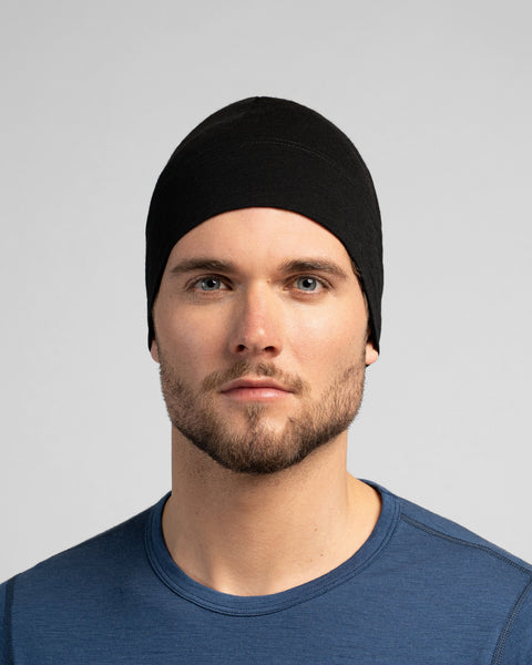 A close-up studio portrait of a man looking directly at the camera. He is wearing a fitted, solid black skull cap and a dark blue crew-neck top. The background is a plain light grey.