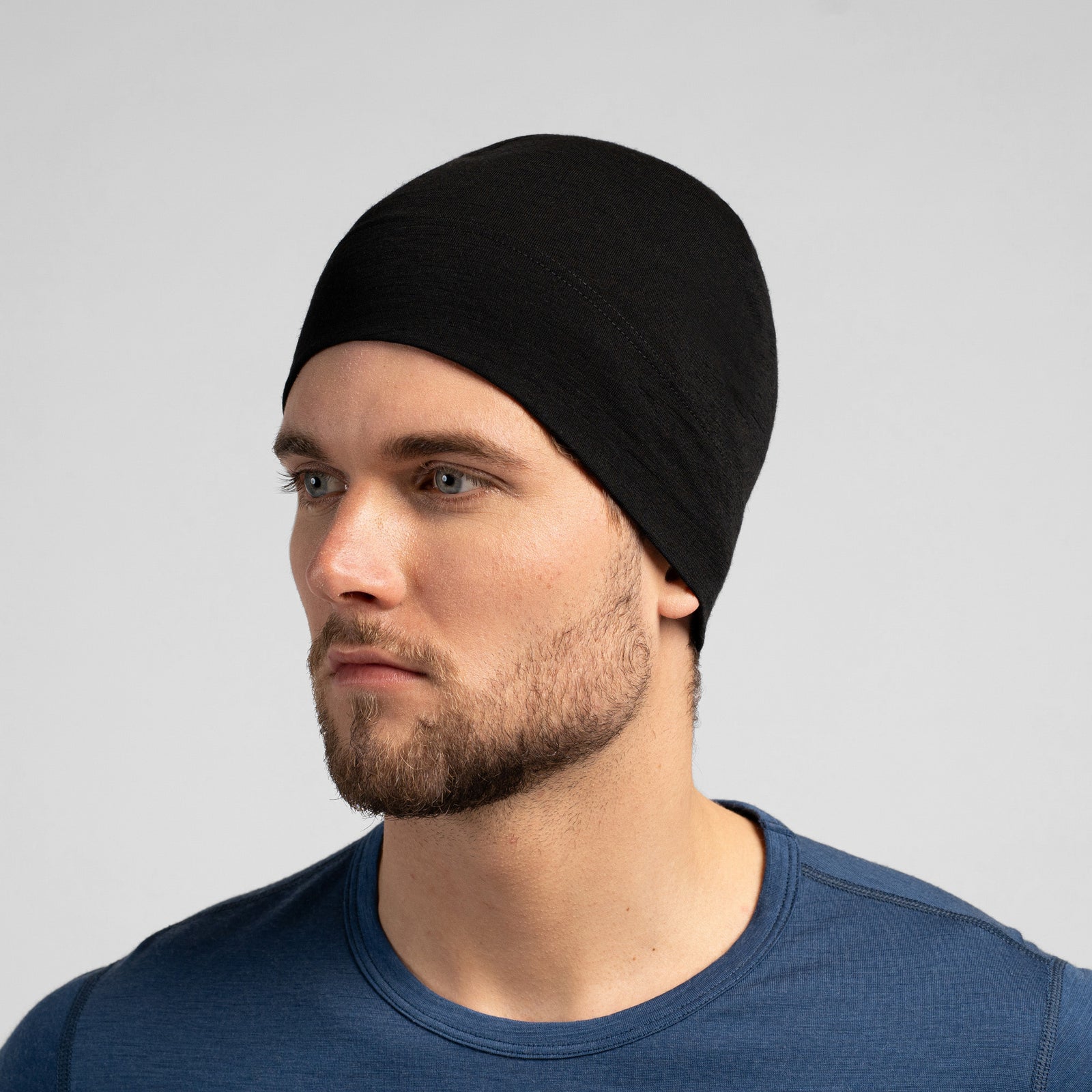 A close-up studio portrait of a man looking thoughtfully off to the left side of the frame. He is wearing a fitted, medium grey skull cap and a solid black crew-neck top. The background is a plain light grey.