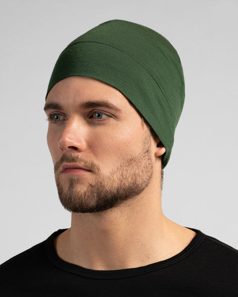 A close-up studio portrait of a man looking thoughtfully off to the left side of the frame. He is wearing a fitted, dark green skull cap and a solid black crew-neck top. The background is a plain light grey.