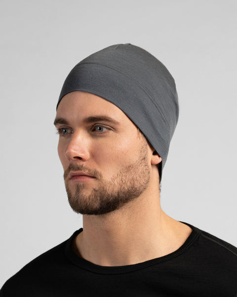 A close-up studio portrait of a man looking thoughtfully off to the left side of the frame. He is wearing a fitted, medium grey skull cap and a solid black crew-neck top. The background is a plain light grey.