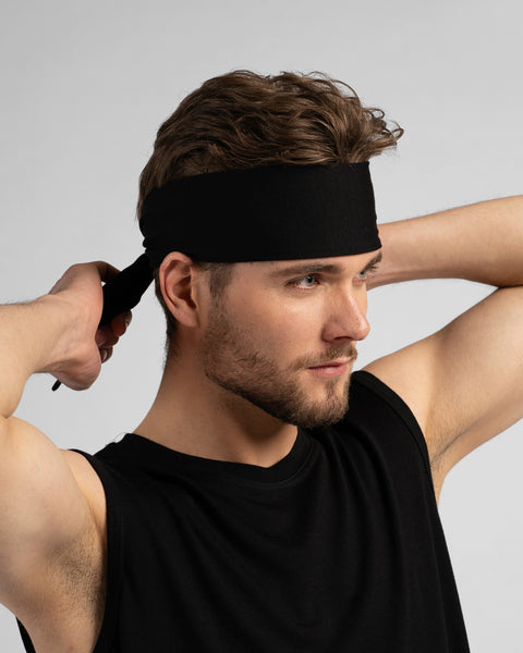 A studio portrait of a man looking downwards and to the right as he ties a solid black headband behind his head. He is wearing a black sleeveless top against a solid light grey background.