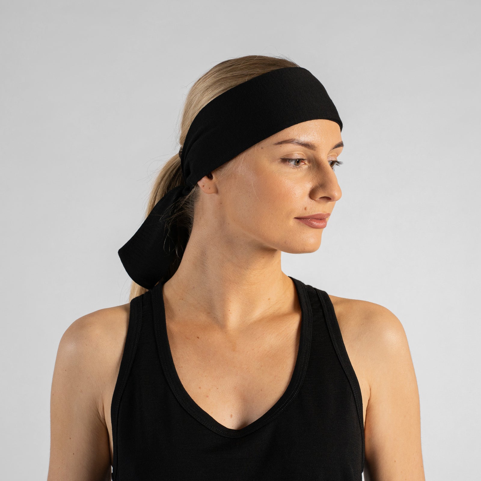 A studio portrait of a woman looking downwards and to the right as she ties a solid black headband behind her head. Her blonde hair is pulled back into a ponytail, and she is wearing a black tank top. The background is a solid light grey.