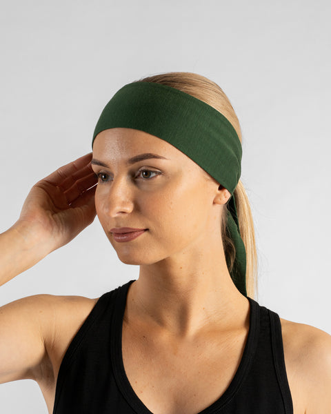 A studio portrait of a woman looking thoughtfully off to the left side of the frame. She is wearing a dark green tie-back headband over her blonde ponytail and a black tank top. Her right hand is lightly touching the side of her head. The background is a solid light grey.