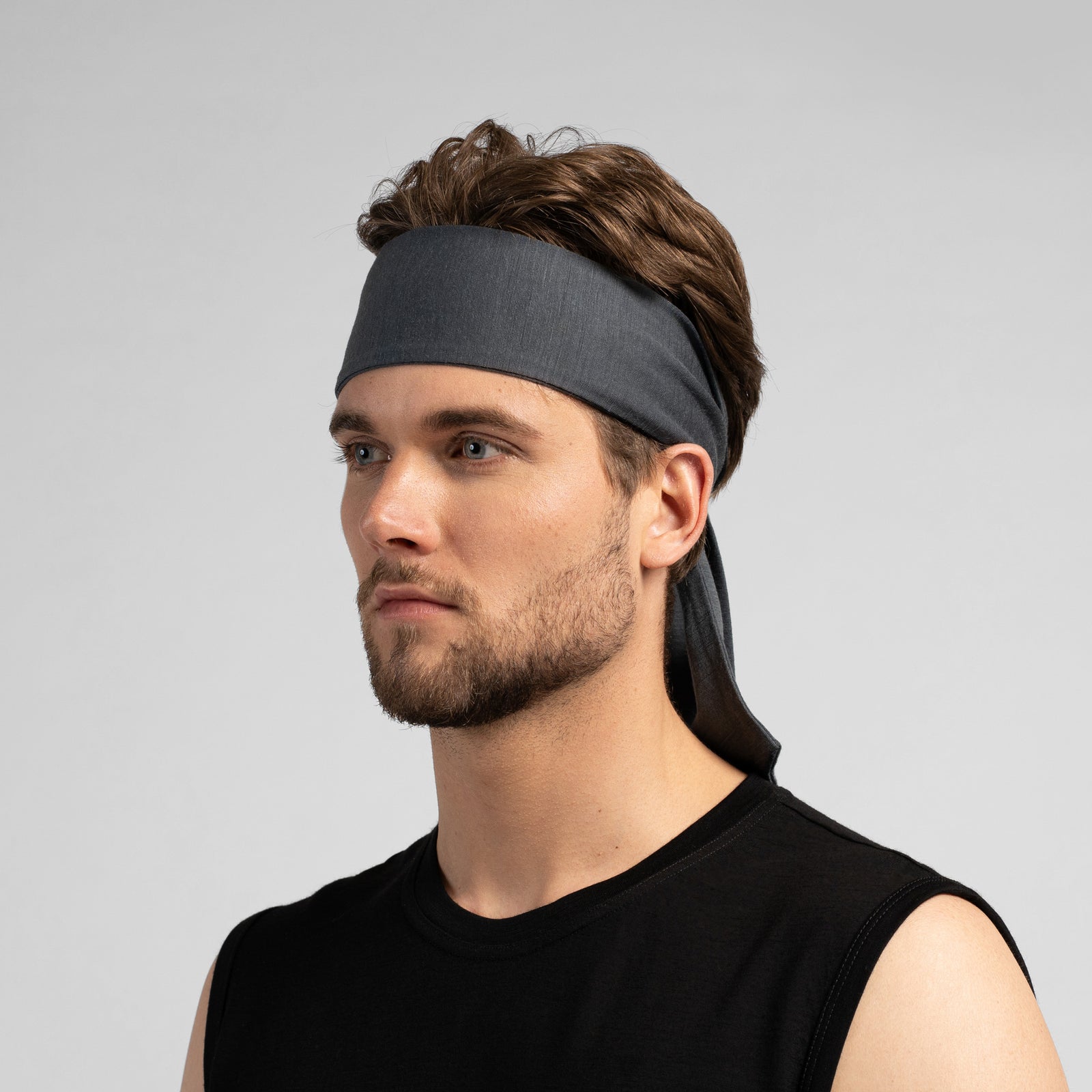 A low-angle shot of a man outdoors tying a blue athletic headband behind his head. He is looking off to the side with a focused expression, wearing a matching blue long-sleeve shirt. The background features a cloudy sky and a blurred, tall stadium light pole.