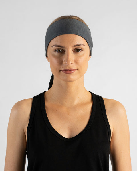 A studio portrait of a blonde woman facing forward and looking directly at the camera against a plain light grey background. She wears a black sleeveless v-neck top and a perfect gray tie headband over a ponytail, with the tail ends hanging down behind her neck.