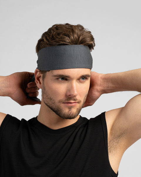 A studio portrait of a man with wavy brown hair looking off to the right while tying a grey headband behind his head. He is wearing a black tank top against a plain light grey background.