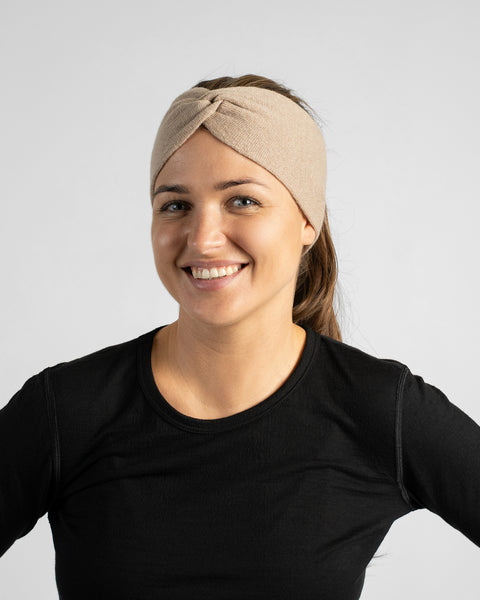 A chest-up portrait of a smiling woman facing forward, wearing a black top and a wide beige knit headband.