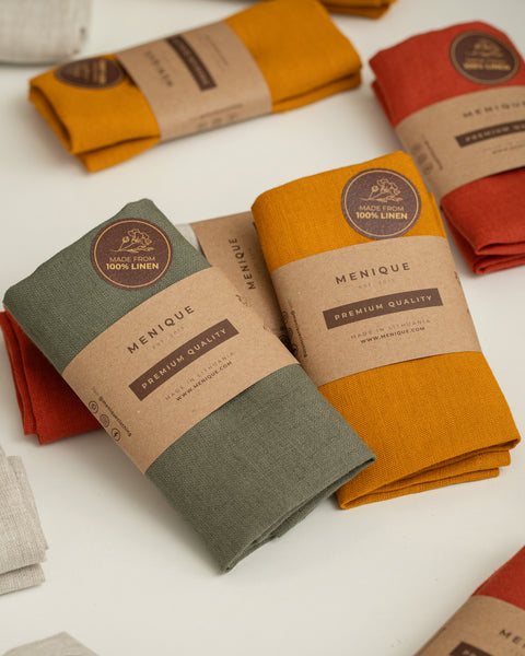 A close-up view of several folded, textured linen napkins in earthy tones like olive green, mustard yellow, rust red, and light beige, scattered on a white surface. The front two napkins are wrapped in brown kraft paper sleeves printed with the brand name "MENIQUE" and feature round brown stickers that read "MADE FROM 100% LINEN."