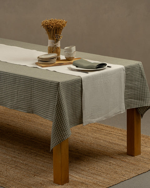 The menique linen tablecloth in green check color, draping over a wooden table, with a vase, wooden board, and a set of dessert plates with tiny clay bowls on top of it.