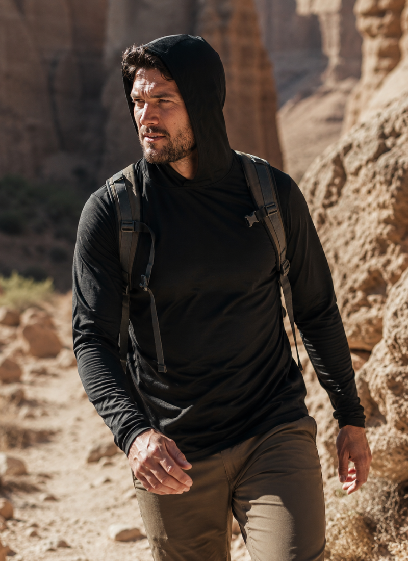 An outdoor shot of a man hiking along a rugged, sunlit canyon trail. He is looking off to the left side of the frame, wearing a black long-sleeved hooded shirt with the hood pulled up, olive green pants, and a backpack.