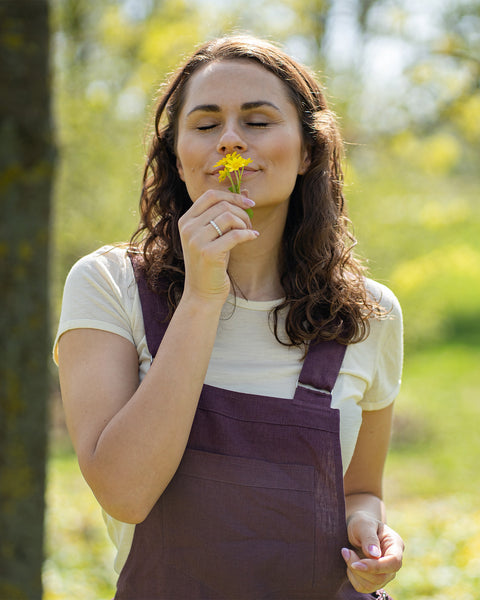 A woman with brown hair, wearing dark purple overalls over a light-colored t-shirt, stands outdoors between two tree trunks on grassy ground.
