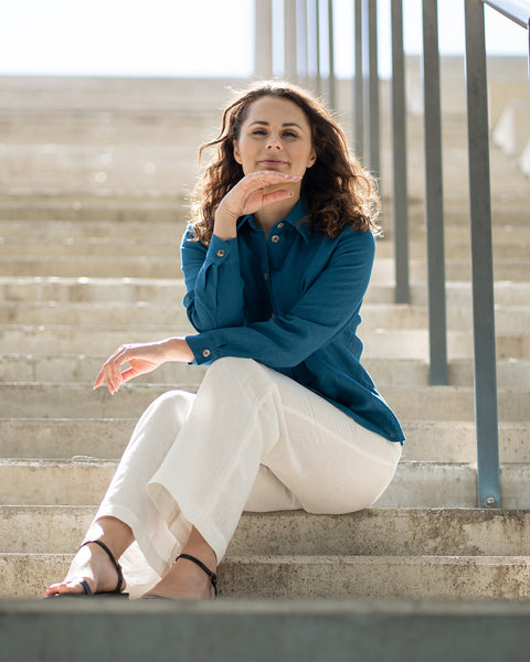 A person with long, dark, curly hair sits casually on light concrete steps. They are wearing a teal long-sleeved collared shirt, white wide-leg pants, and dark sandals. Their right hand is under their chin, and they are looking towards the viewer with a slight smile. A metal railing is visible on the right side of the frame.