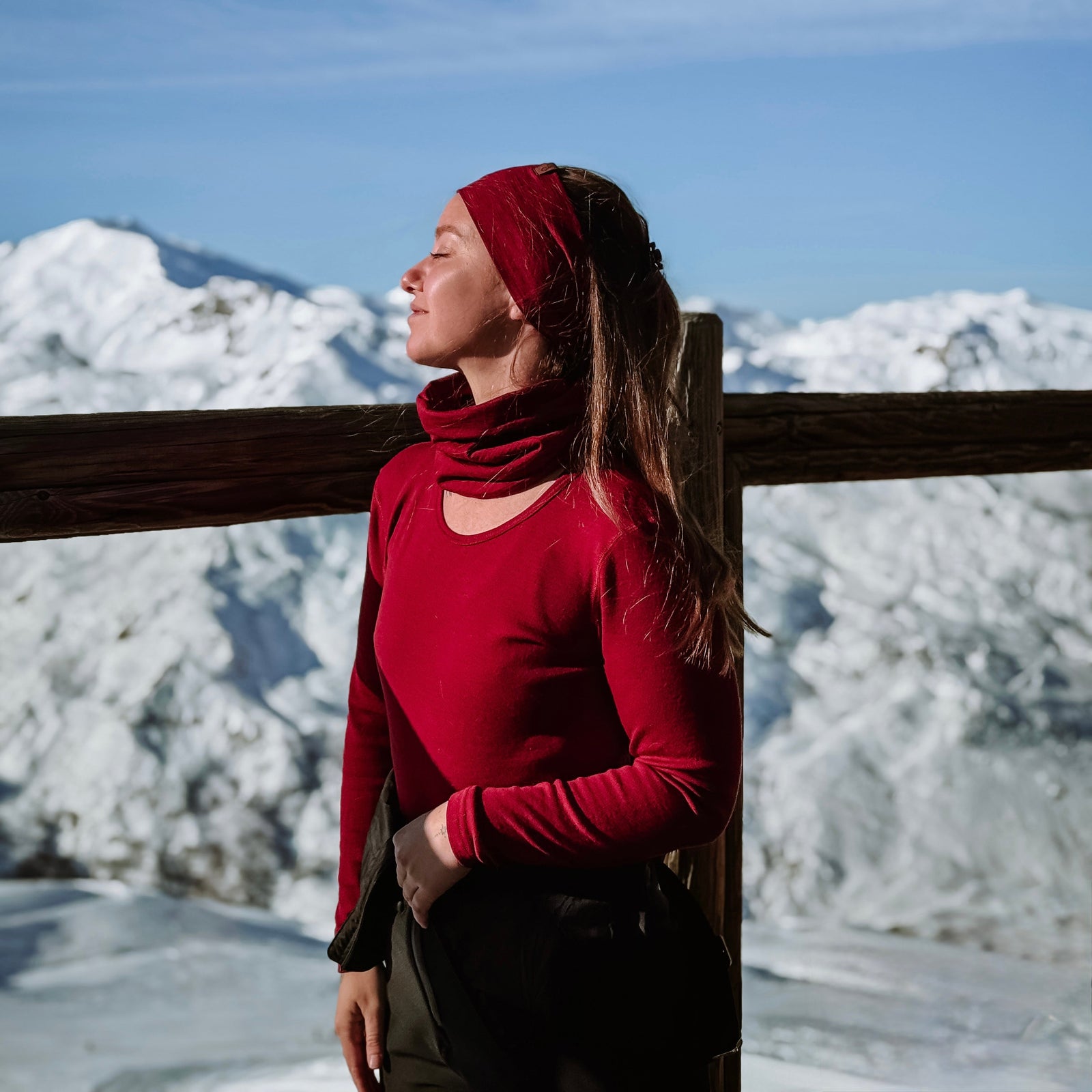 Woman in red outfit standing by a wooden railing with snow-covered landscape and blue sky in the background