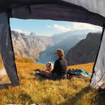 A view from inside a tent looking out at a person sitting on a grassy hillside with a small dog. They are looking out over a spectacular fjord or mountain valley landscape. The dark edges of the tent opening frame the scenic view.