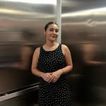 A slightly blurry photo of a woman standing inside an elevator with reflective metal walls. She is wearing a black sleeveless dress with white polka dots and has her hands clasped in front of her.