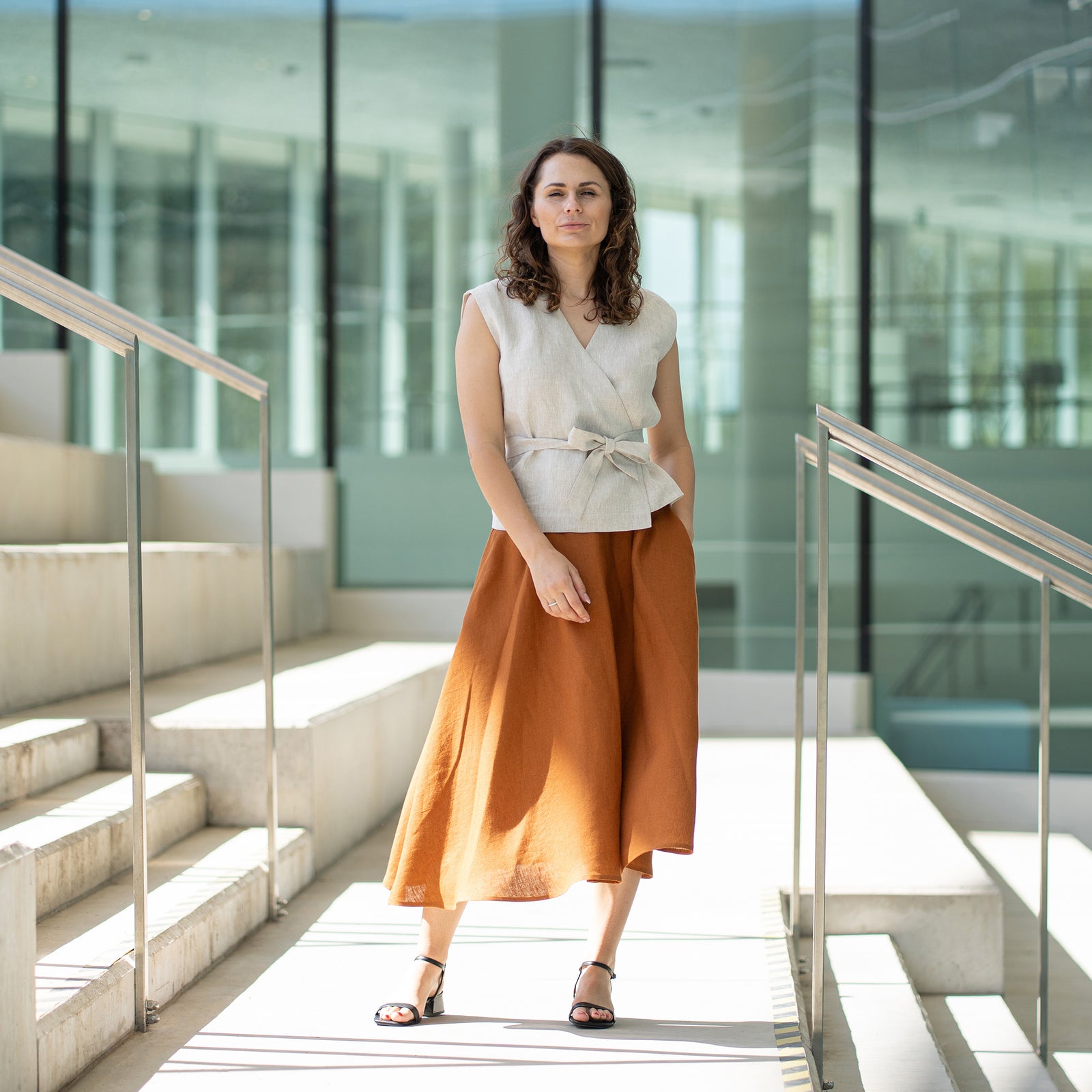 A woman with brown hair, wearing a light beige wrap top and a long rust-brown skirt, poses indoors in front of large windows.

