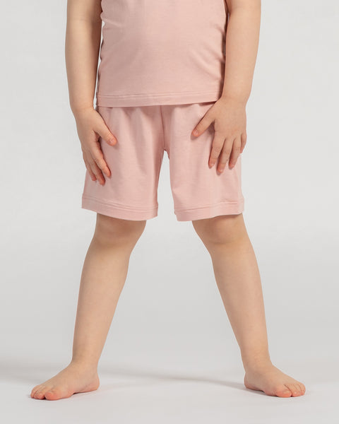 A studio shot, cropped from the chest down, of a young child standing barefoot. The child has their hands resting lightly on the front of their thighs. They are wearing a matching set of a light dusty pink short-sleeved t-shirt and shorts. The background is a plain light grey.