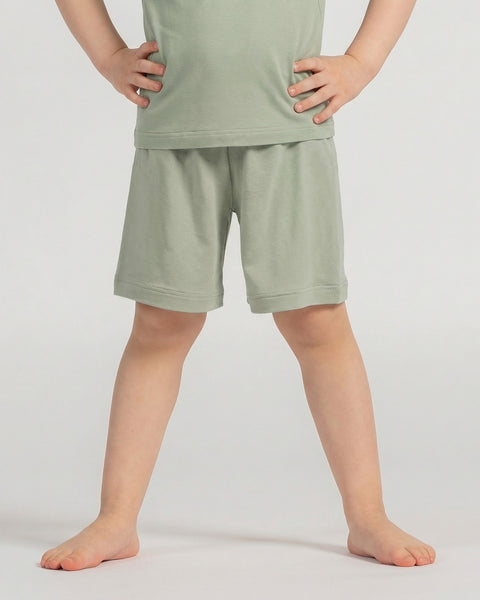 A studio shot, cropped from the chest down, of a young child standing barefoot. The child has their hands resting on their hips. They are wearing a matching set of a light sage green short-sleeved t-shirt and shorts. The background is a plain light grey.