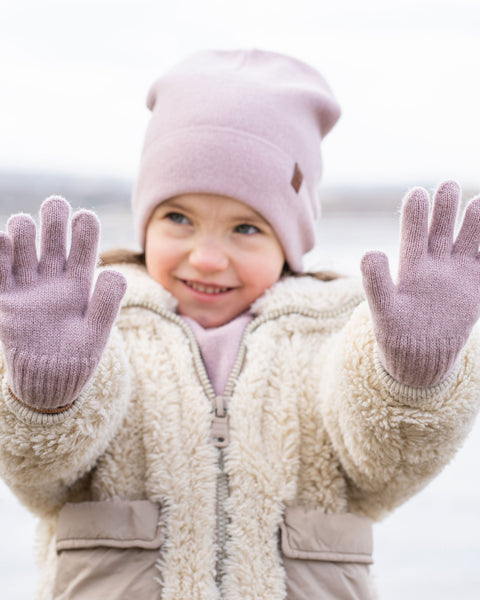 A smiling young girl holding both hands open towards the camera, showing off light dusty pink knitted gloves. She is wearing a matching light pink beanie, a matching neck warmer, and a cozy cream-colored fleece jacket. The background is a softly blurred outdoor winter scene.