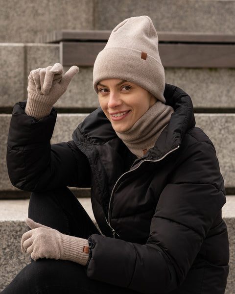 Woman sitting on outdoor stone steps, wearing a beige knit beanie, matching gloves, and neck warmer with a black puffer jacket, smiling and holding up one hand in a casual pose.