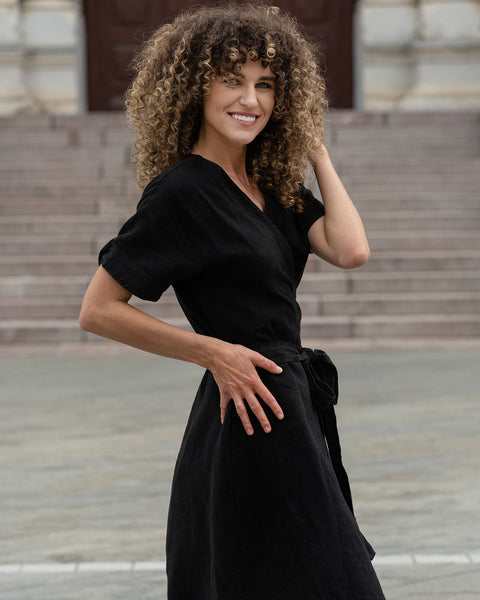 A person with medium-length curly hair smiles and poses, wearing a black, short-sleeved wrap dress with a tie at the waist. They are standing in front of a building with a large stone staircase in the background. Their hand is resting on their hip.