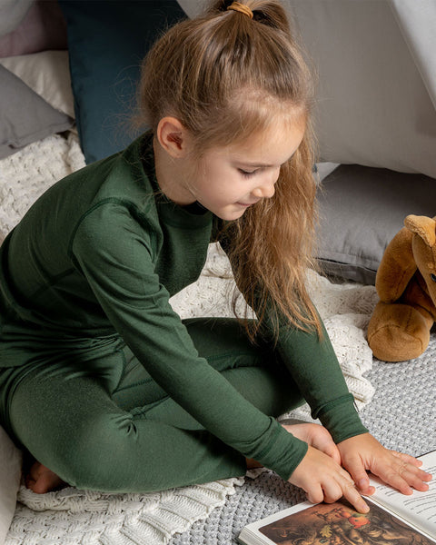 A young girl sits comfortably on a textured white blanket surrounded by cozy pillows, wearing a dark green long-sleeve top and matching leggings. She is attentively pointing at an illustration in an open book, with a brown plush teddy bear resting beside her.
