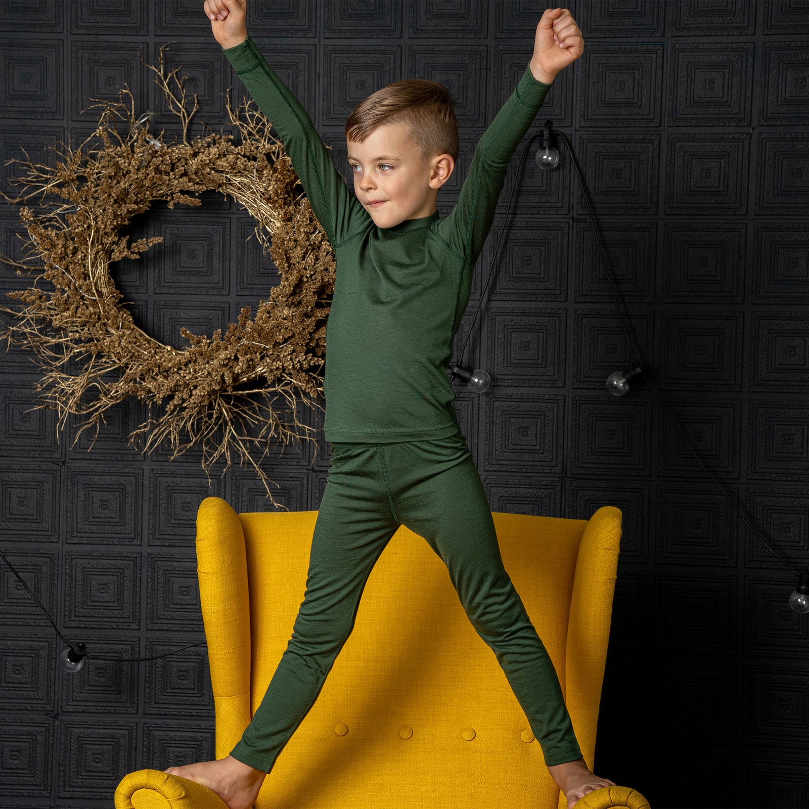 A boy sitting an a chair, with one leg on top of it, wearing the menique Kids' 160 Merino Pants in Dark Green color. He is resting his head on the raised leg's knee.