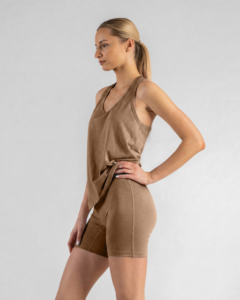 A model in a side-profile view poses against a light gray background, wearing a matching beige tank top and bike shorts, with her right hand resting on her hip.
