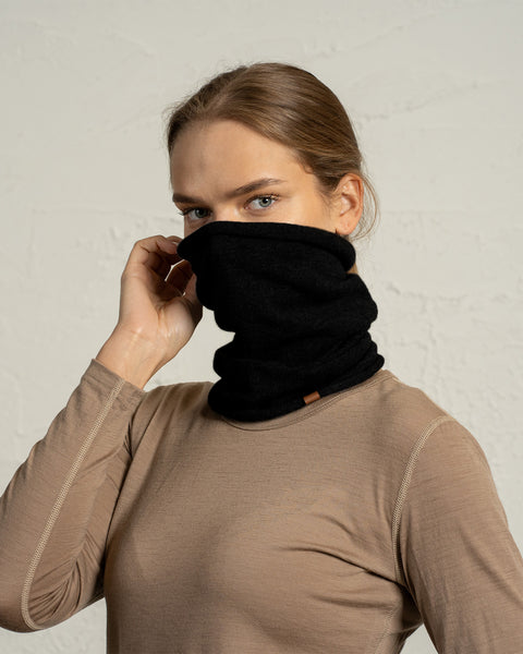 A woman wearing a beige long-sleeve top holds a black wool snood neck gaiter over the lower half of her face, covering her mouth and nose. Her blue eyes look directly at the camera, and her hair is pulled back in a neat, low style. The textured light-colored background keeps the focus on the gaiter and her expression.