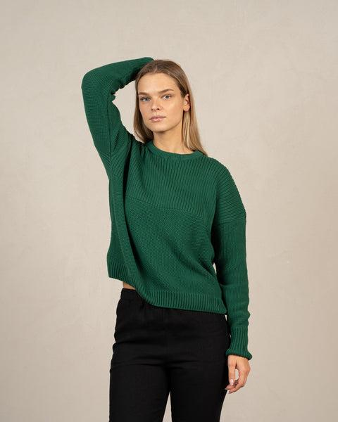 A young blond woman wearing the mennique.combed cotton sweater SONATA in green color, with one of her hands behind her head.