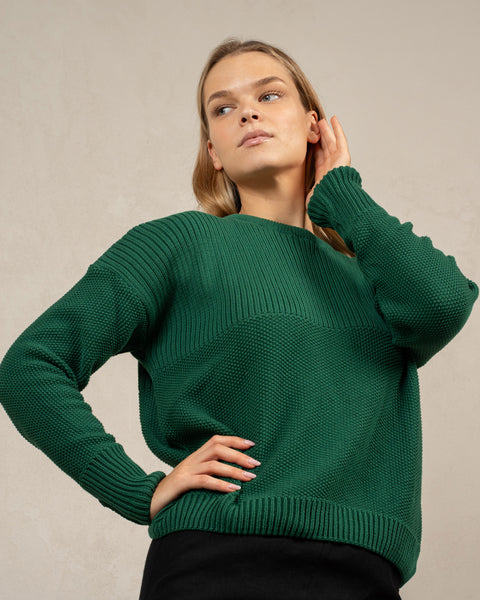 A woman captured from below at an angle, wearing the mennique.combed cotton sweater in green color. She has one of her hands on her hip, and the other is brushing lightly against the side of her face. The sweater is knitted in a honeycomb stitched.