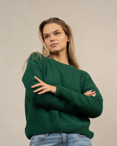 A woman captured from an angle from below, wearing the mennique.combed cotton sweater SONATA in green color. She has her arms crossed over her waist.