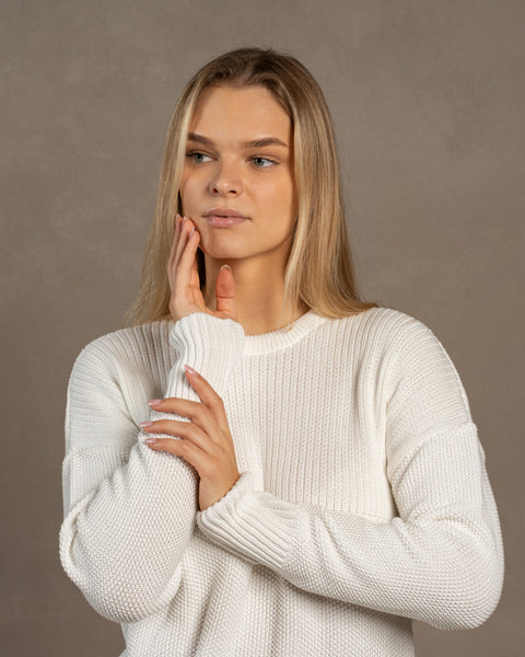 A woman captured wearing the mennique.combed cotton sweater in white color. She has both of her hands raised near her face. The sweater is knitted in a honeycomb stitched.