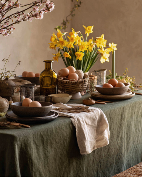 A rustic, moody table setting anchored by a textured stone green linen tablecloth. Dark, speckled ceramic bowls and plates are arranged alongside a woven basket filled with brown eggs, while a large bouquet of bright yellow daffodils and a slim green taper candle provide a pop of seasonal color.